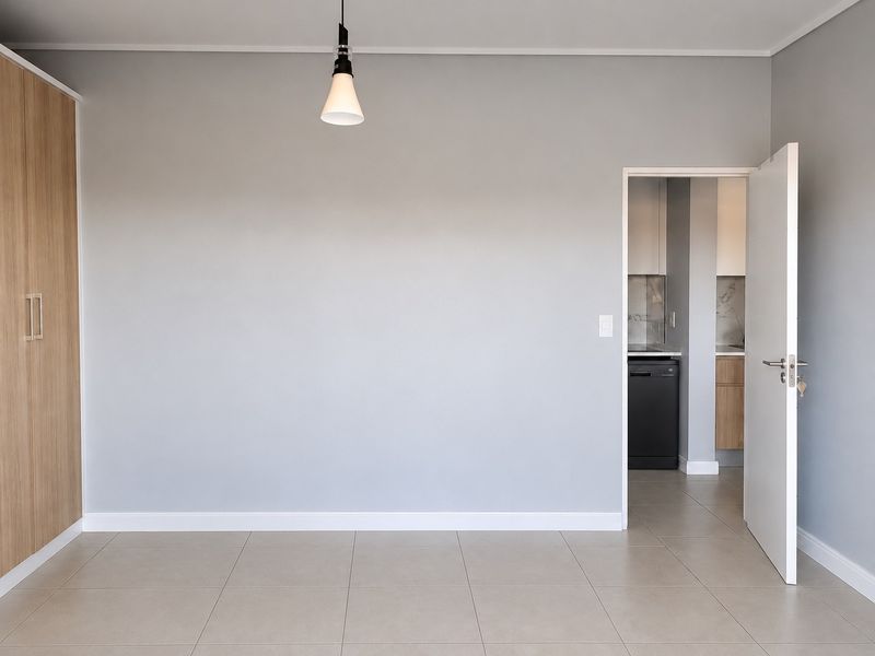 A bright, empty bedroom featuring neutral tiled flooring, a modern built-in wardrobe, and a clean, minimalist aesthetic with an adjacent view into the kitchen area.