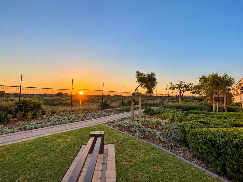 A well-maintained landscaped garden area featuring a paved walkway, manicured lawn, and secure perimeter fencing at sunset.