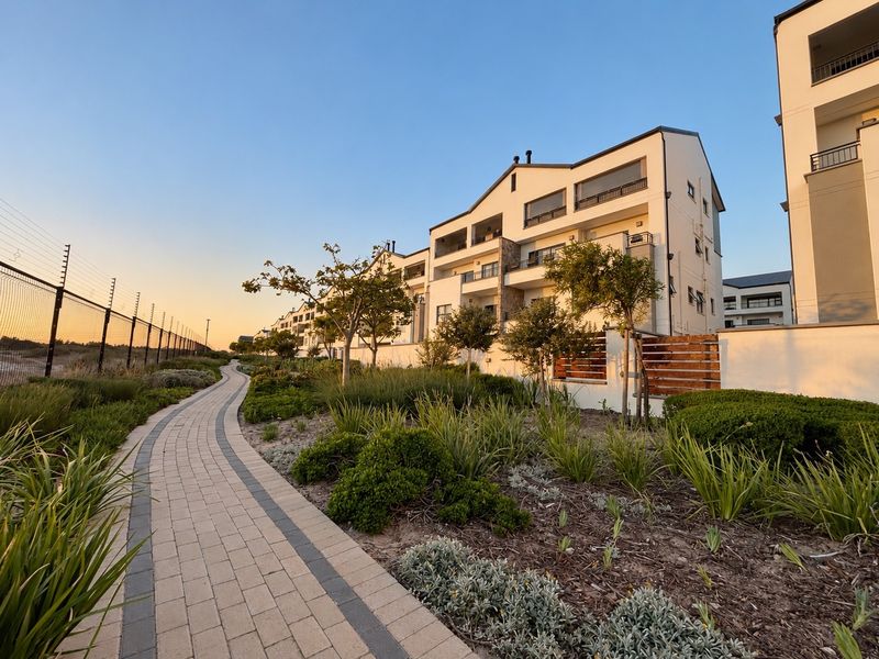 A well-maintained apartment complex exterior featuring a paved walkway, landscaped gardens, and secure perimeter fencing at sunset.