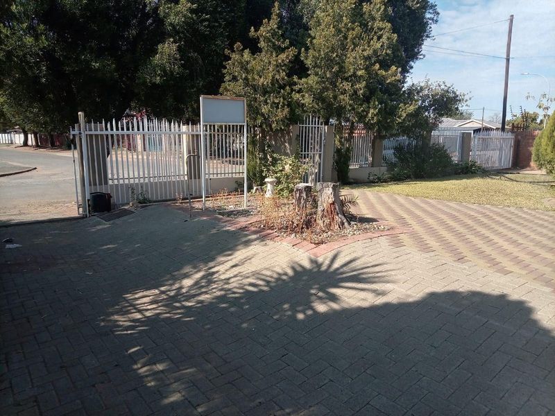 Front exterior view featuring a paved driveway, metal security gate, and perimeter fencing with established garden areas.