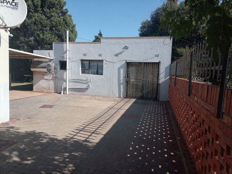 Rear exterior view showing a paved driveway, brick boundary wall, and a small outbuilding with security gates and bars on the windows.