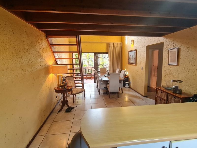 Open-plan living and dining area featuring exposed timber ceiling beams, tiled flooring, and a staircase leading to an upper level with access to a patio.