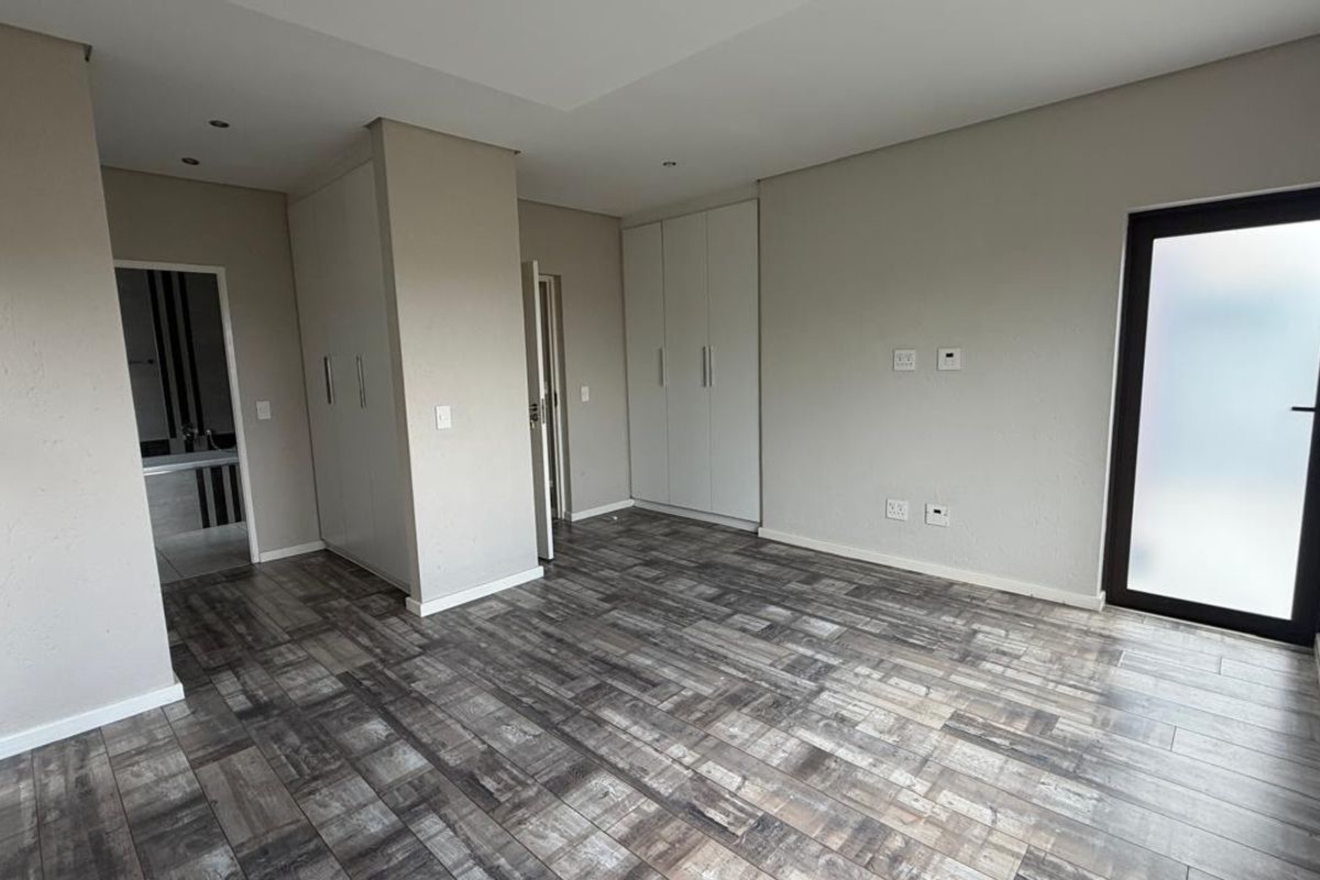 A spacious, modern bedroom featuring neutral wall tones, wood-look tiled flooring, ample built-in cupboard space, and a private frosted glass door.