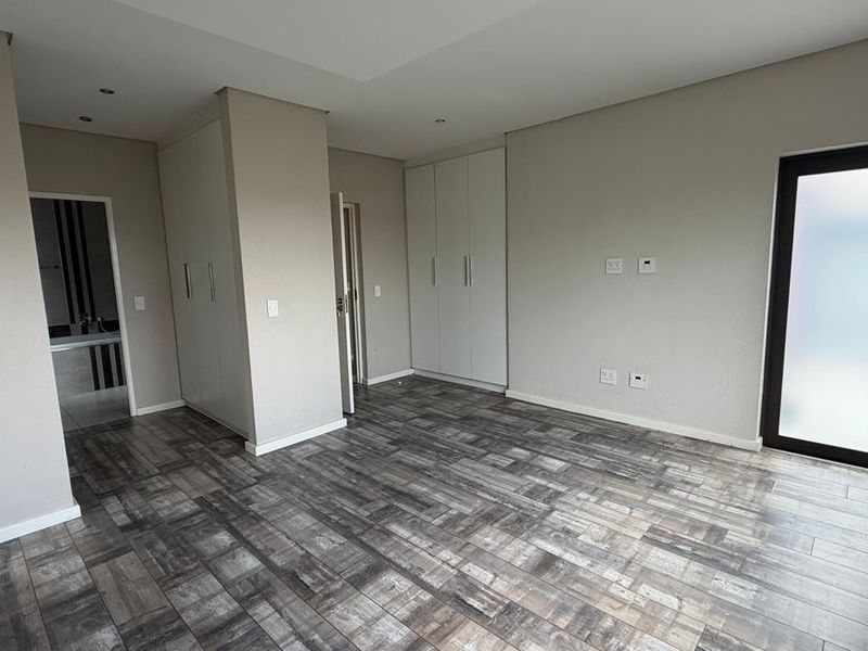 A spacious, modern bedroom featuring neutral wall tones, wood-look tiled flooring, ample built-in cupboard space, and a private frosted glass door.