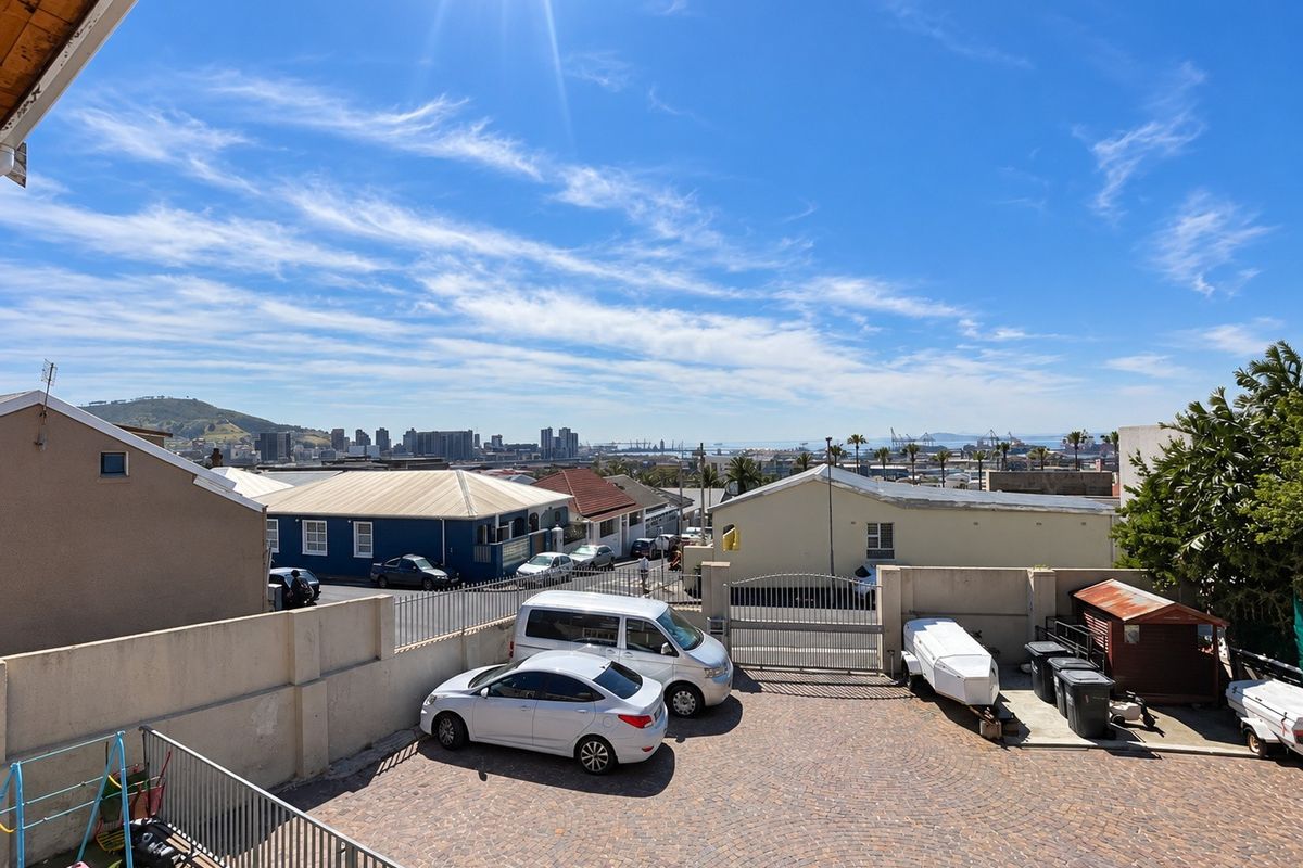 An elevated view of the rear paved driveway and parking area, featuring perimeter walling, security gates, and a distant cityscape view.