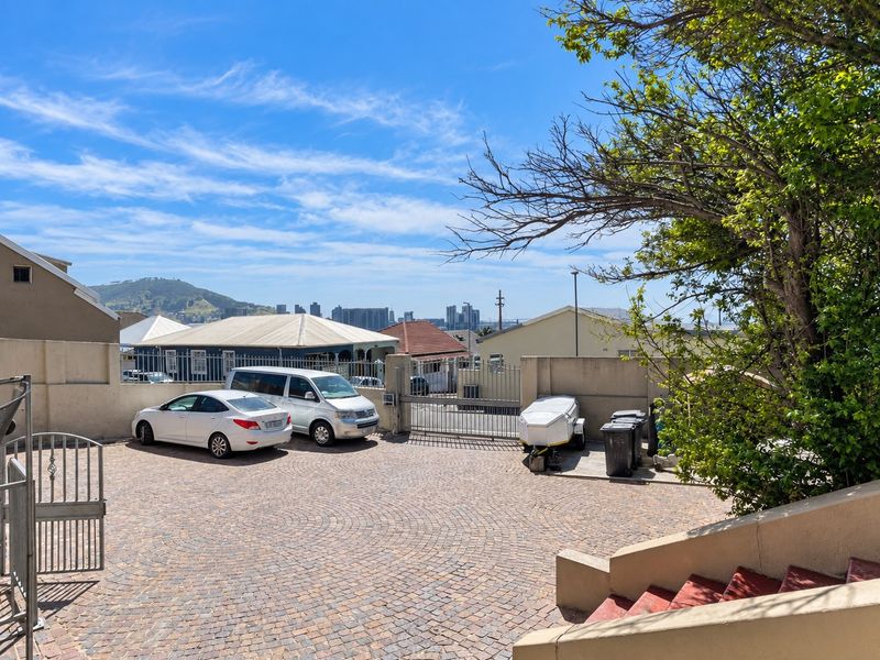 A paved driveway area featuring a secure metal gate, perimeter walling, and concrete stairs, offering a view of the surrounding neighborhood and distant city skyline.
