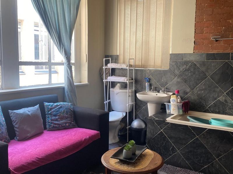 A bathroom featuring a built-in bathtub with slate tiling, a pedestal basin, and toilet, accented by an exposed brick wall and large window.