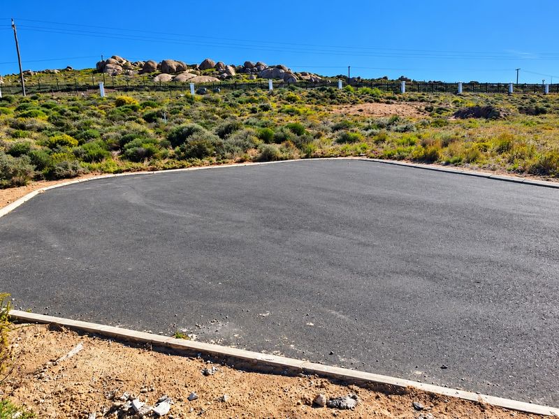 A paved asphalt driveway area on a vacant residential plot, featuring concrete curbing and perimeter fencing against a natural rocky landscape.