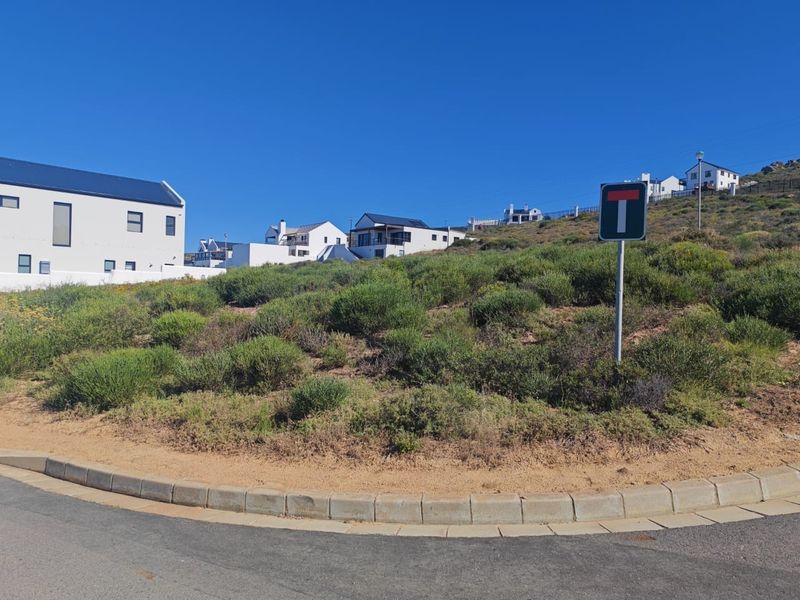 A vacant residential plot situated at the end of a cul-de-sac, featuring natural vegetation and established kerbing in a developed coastal estate.