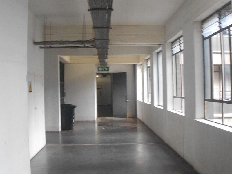 A long, industrial-style hallway featuring exposed cable trays, concrete flooring, and large windows providing natural light.