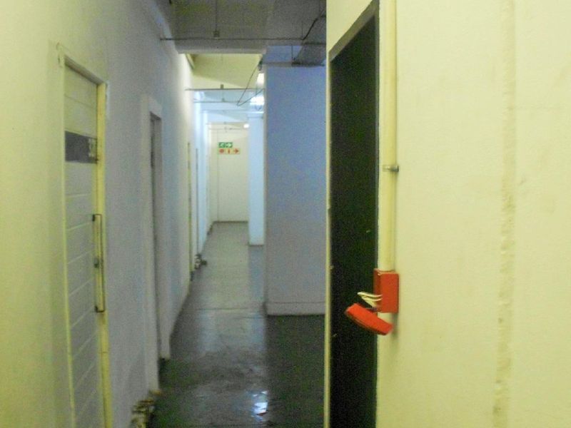 A utilitarian apartment hallway featuring exposed electrical conduits and a damaged fire alarm call point, showing signs of significant wear and maintenance requirements.