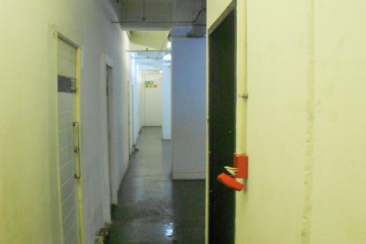 A utilitarian apartment hallway featuring exposed electrical conduits and a damaged fire alarm call point, showing signs of significant wear and maintenance requirements.