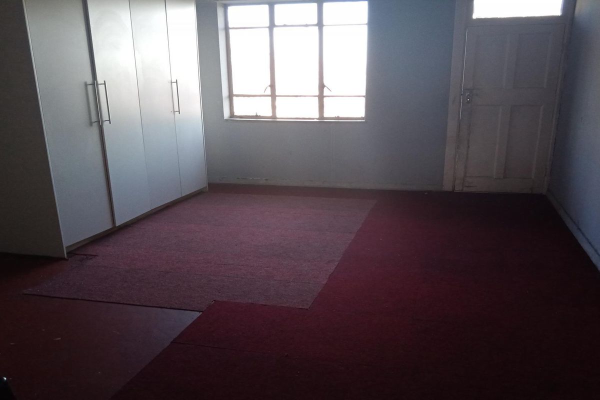 An empty bedroom featuring built-in cupboards, a multi-pane window, and worn carpet flooring in need of replacement.