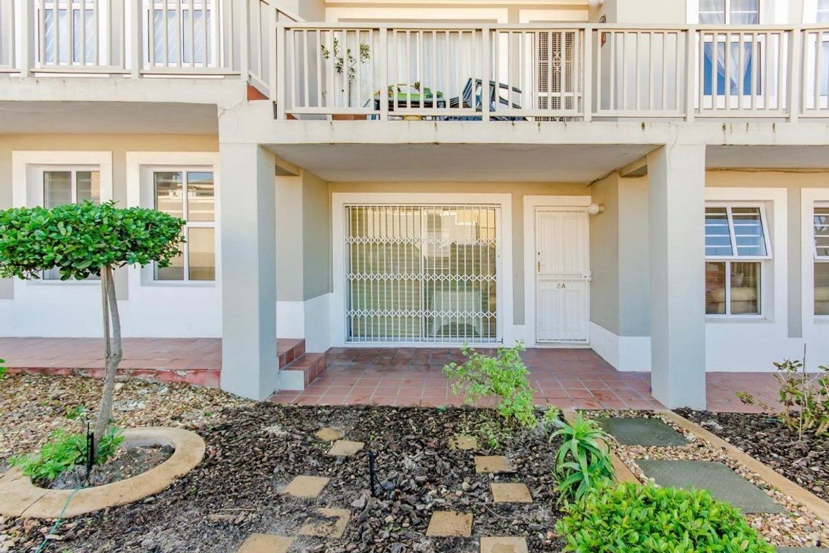 Ground floor patio area with tiled flooring, security gate access, and an overhead balcony, set within a landscaped garden environment.