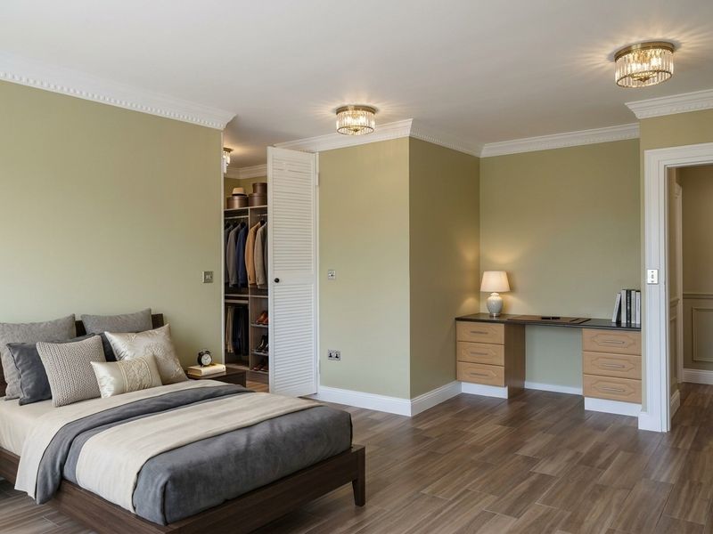 Spacious bedroom featuring sage green walls, wood-look flooring, crown molding, a built-in desk area, and a walk-in wardrobe with louvered doors.
