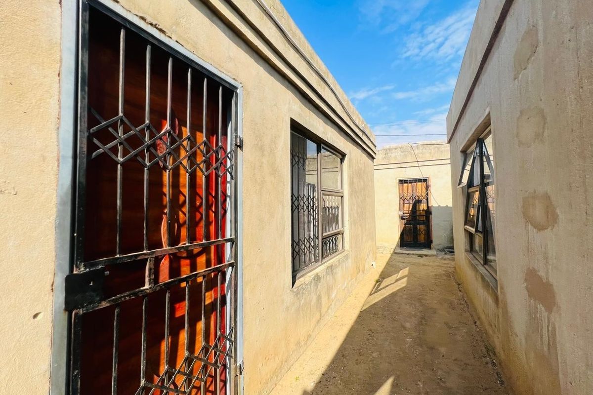 A narrow exterior side passage between two structures featuring security gates and metal-framed windows with security bars.