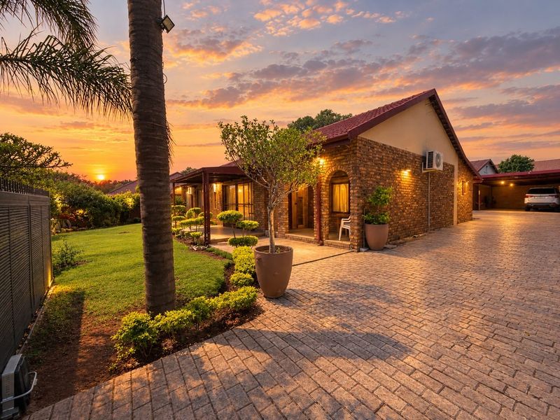 A well-maintained brick house featuring a paved driveway, covered patio, and landscaped garden, captured during a beautiful sunset.