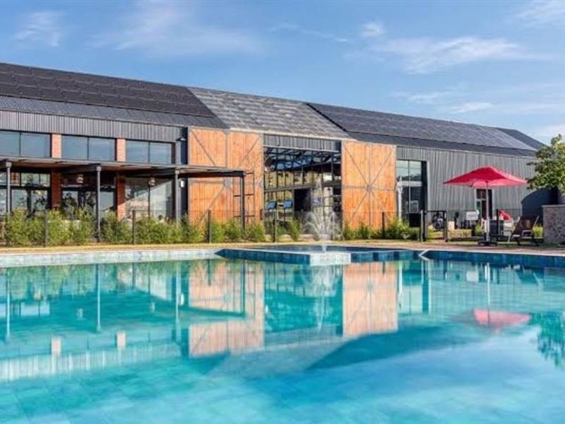 A modern clubhouse or recreational facility featuring a large swimming pool with a water fountain, set against a contemporary building with solar panels.