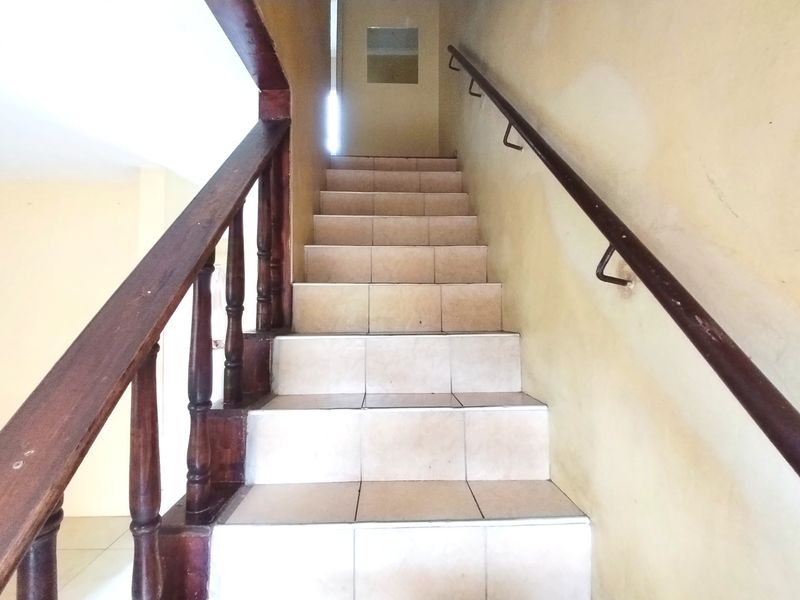 A staircase featuring tiled steps, a wooden balustrade, and a wall-mounted handrail, showing signs of wear on the surrounding wall surfaces.