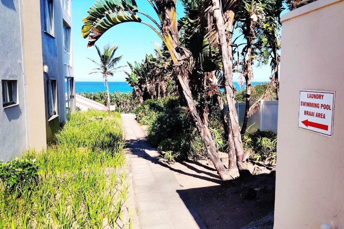 A paved walkway leading through a landscaped garden area between apartment buildings, featuring mature trees and a view towards the ocean.