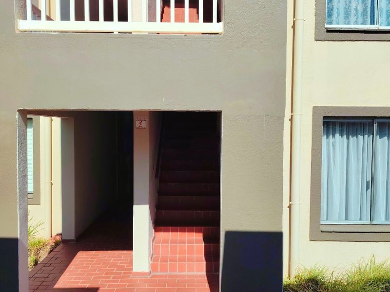Exterior view of an apartment building entrance featuring a tiled staircase, covered walkway, and balcony with white railings.