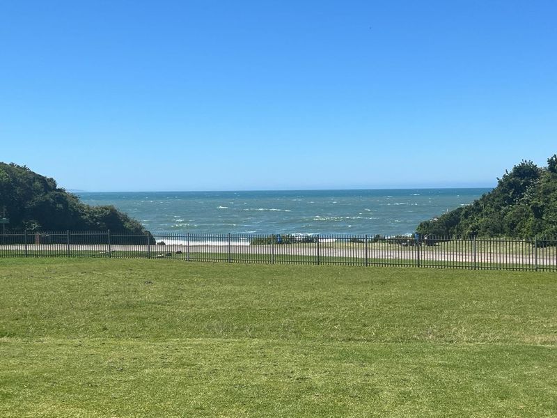 A scenic view of the ocean framed by coastal vegetation, featuring a well-maintained lawn and a secure metal perimeter fence.