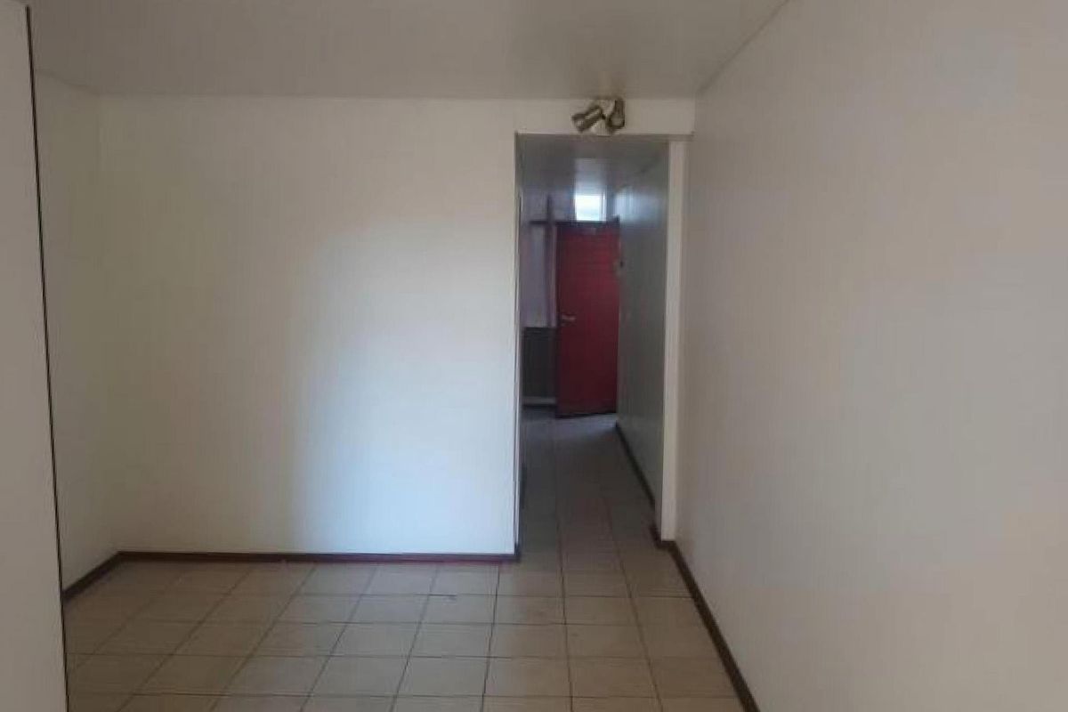 An empty living area featuring tiled flooring and a simple spotlight fixture, leading into a hallway with a red door.