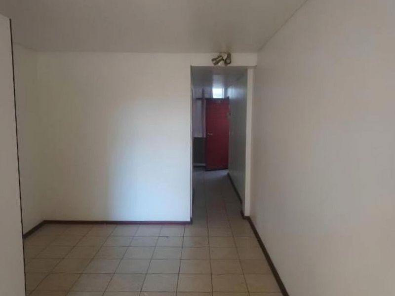 An empty living area featuring tiled flooring and a simple spotlight fixture, leading into a hallway with a red door.