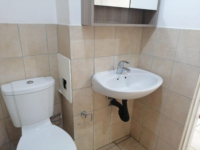 A functional bathroom featuring a wall-mounted basin, toilet, and tiled finishes, currently requiring minor repairs to wall tiling and plumbing aesthetics.