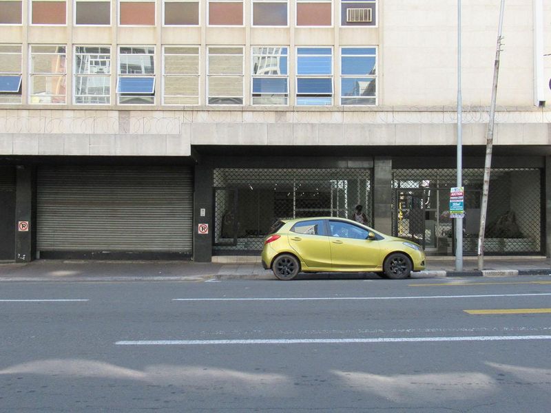 Street-level view of a commercial or mixed-use building facade featuring security roller shutters, metal gates, and large display windows.