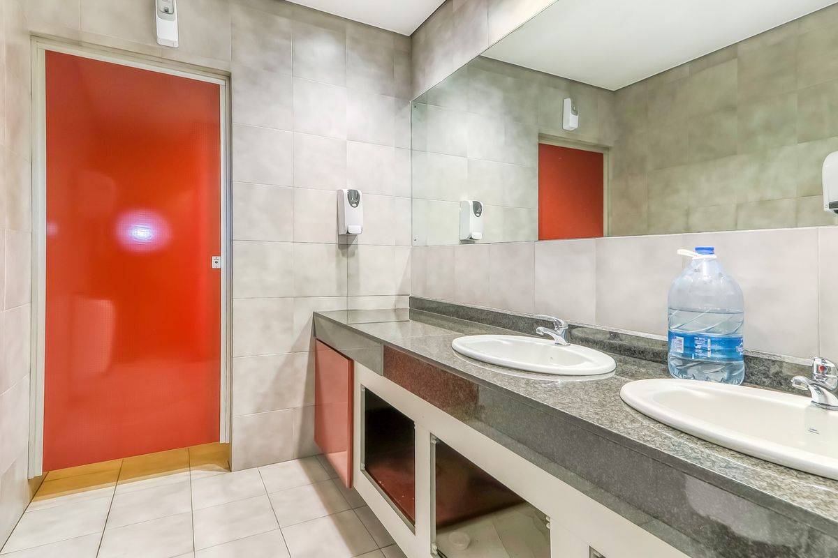 A functional office bathroom featuring a dual vanity with granite countertops, large wall mirror, and tiled finishes, currently requiring minor cabinet repairs.