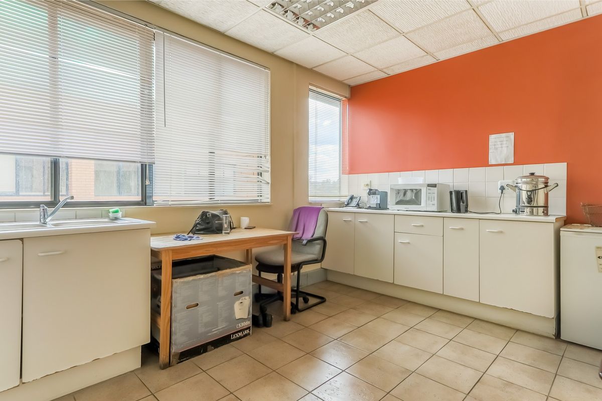 A functional office kitchenette featuring built-in cabinetry, tiled backsplash, stainless steel sink, and tiled flooring with ample natural light from large windows.