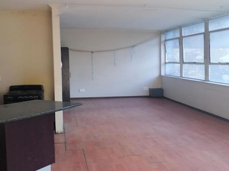 An open-plan living and kitchen area featuring laminate flooring, large windows, and a functional kitchen layout, currently vacant and ready for renovation.