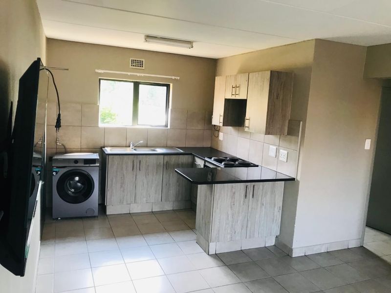 A functional kitchen featuring light wood-look cabinetry, dark countertops, a built-in electric hob, and tiled flooring with an integrated laundry appliance space.