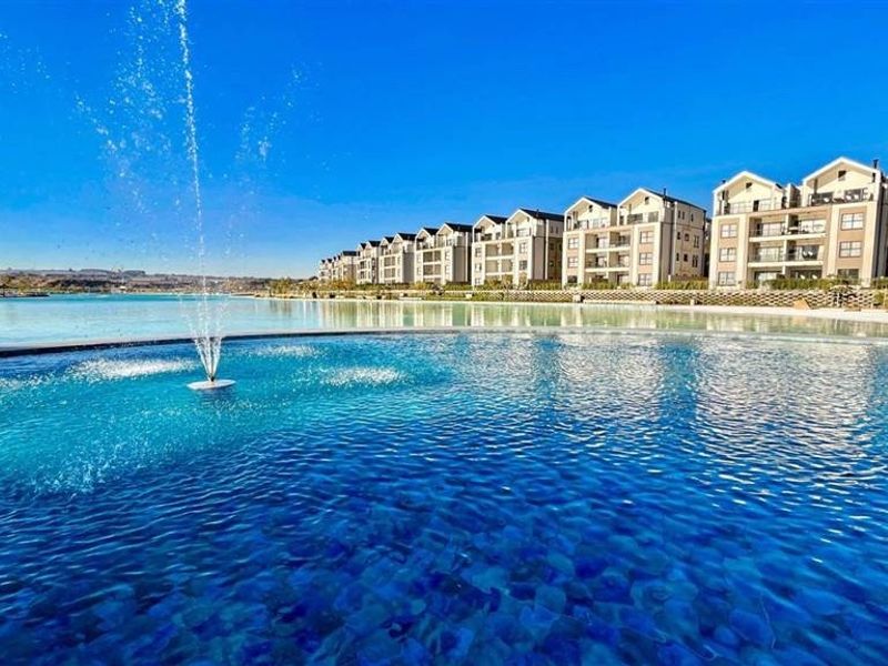 A scenic view of a large, clear swimming pool with a central water fountain, set against a backdrop of modern apartment buildings.