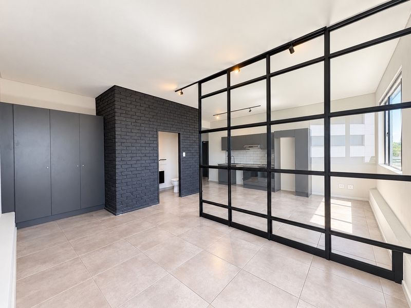 Modern open-plan living area featuring a striking black steel-framed glass partition, exposed brick wall, and sleek kitchen cabinetry with integrated appliances.