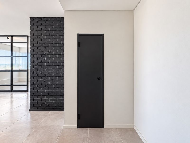A modern hallway featuring a striking black painted brick wall, sleek tiled flooring, and a black framed glass partition leading to an adjacent room.