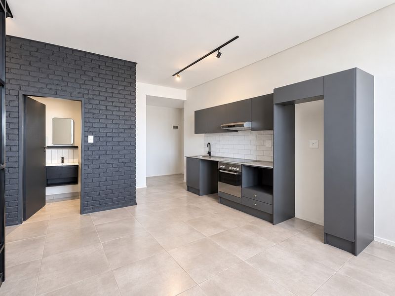 Modern open-plan kitchen featuring sleek dark cabinetry, built-in oven, extractor fan, and a stylish exposed brick feature wall with neutral tiled flooring.