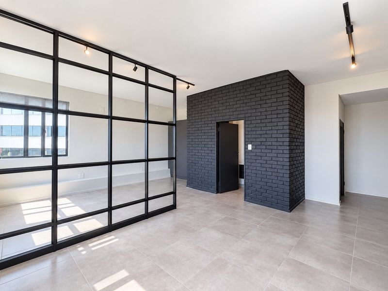 Modern open-plan living area featuring a striking black metal-framed glass partition, exposed black brick feature wall, and contemporary track lighting on tiled floors.