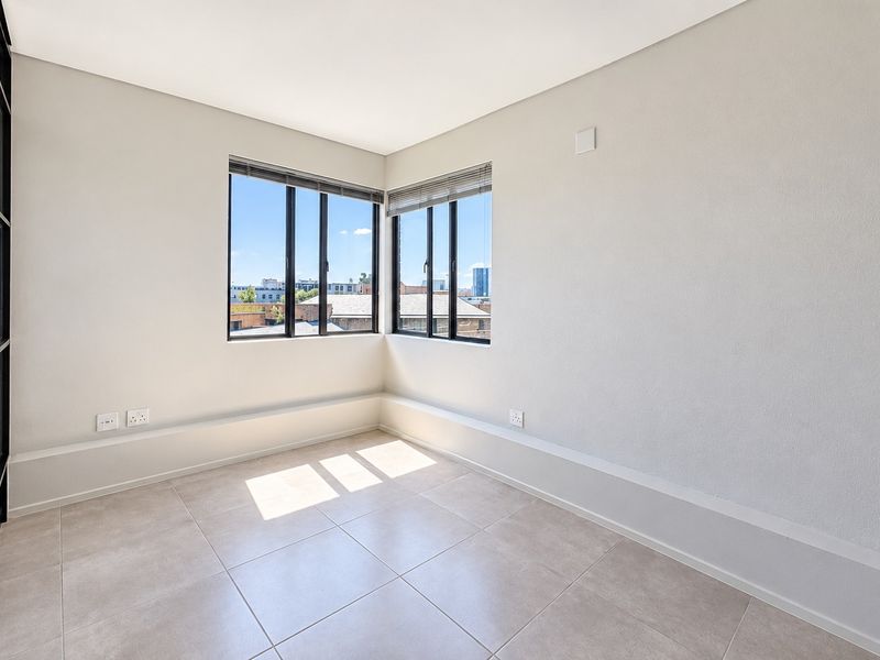 A bright, empty bedroom featuring modern tiled flooring, corner windows with black frames, and neutral wall finishes, offering a clean and versatile living space.