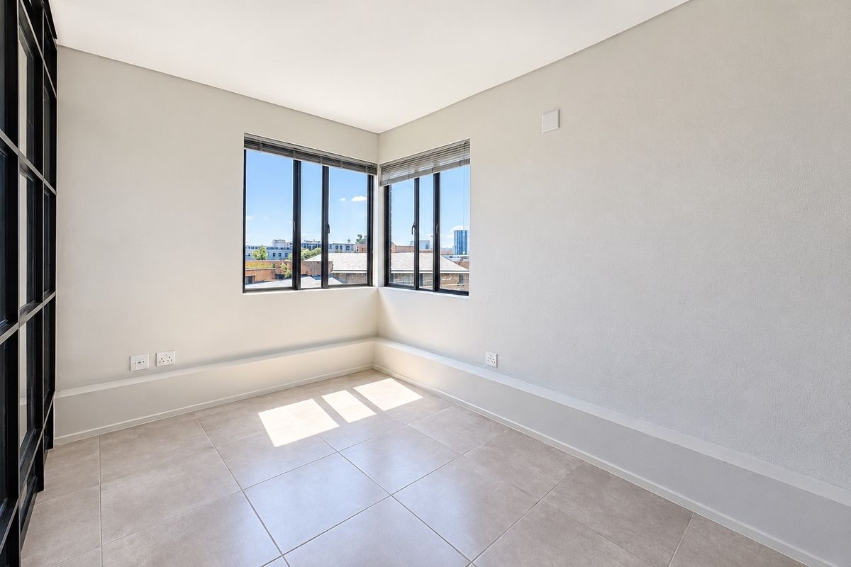A bright, empty bedroom featuring modern tiled flooring, corner windows with black frames, and neutral wall finishes, offering a clean and versatile living space.