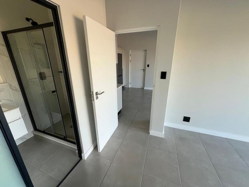 A modern, empty bedroom featuring neutral tiled flooring, clean white walls, and an adjoining en-suite bathroom with a contemporary black-framed shower enclosure.