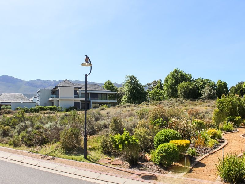 A street-level view of a residential plot featuring a paved walkway, established garden landscaping, and a street lamp, with neighboring properties and mountain views.