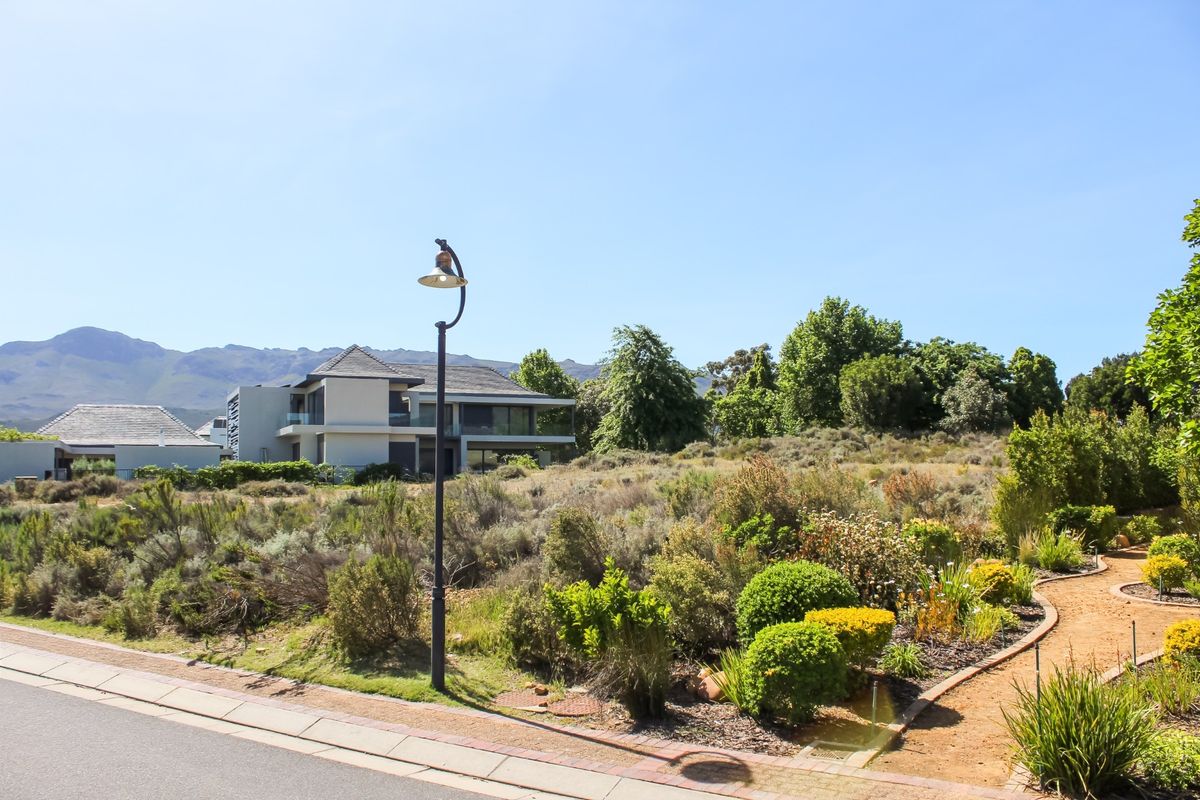 A street-level view of a residential plot featuring a paved walkway, established garden landscaping, and a street lamp, with neighboring properties and mountain views.