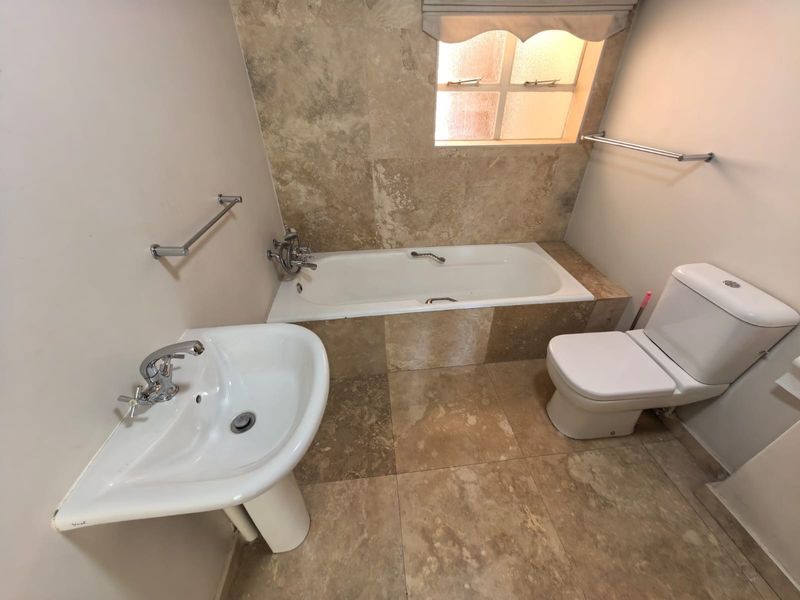 A clean bathroom featuring a built-in bathtub with travertine tiling, a pedestal basin, and a standard toilet, finished with chrome fixtures.