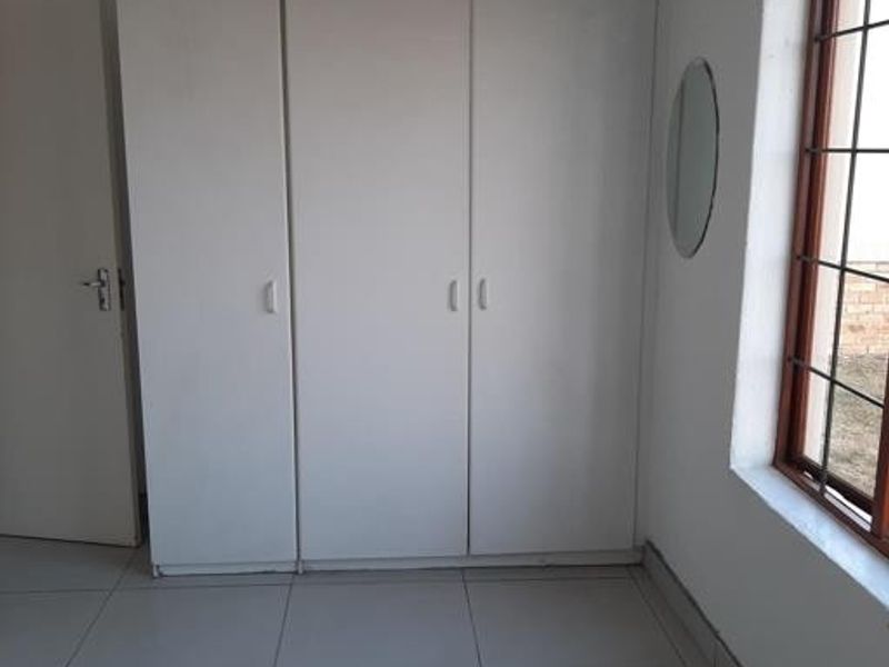 A clean, empty bedroom featuring white built-in cupboards, neutral tiled flooring, and a window with security bars.