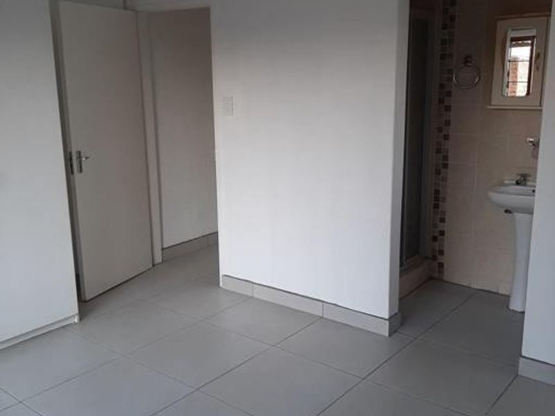 A clean, empty bedroom featuring neutral tiled flooring, a built-in cupboard, and direct access to an en-suite bathroom with a pedestal basin and shower.