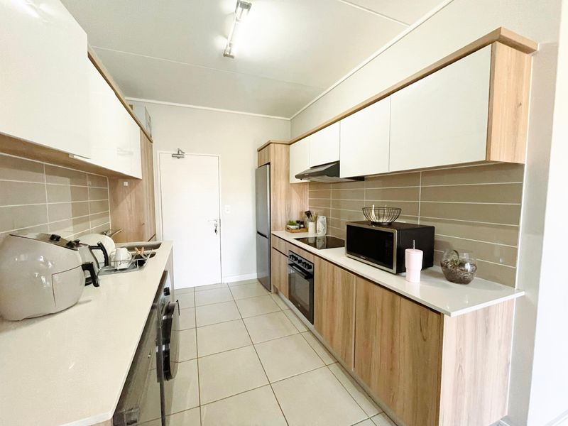 Modern galley-style kitchen featuring light wood-grain cabinetry, white countertops, tiled backsplash, and integrated appliances including an oven and hob.