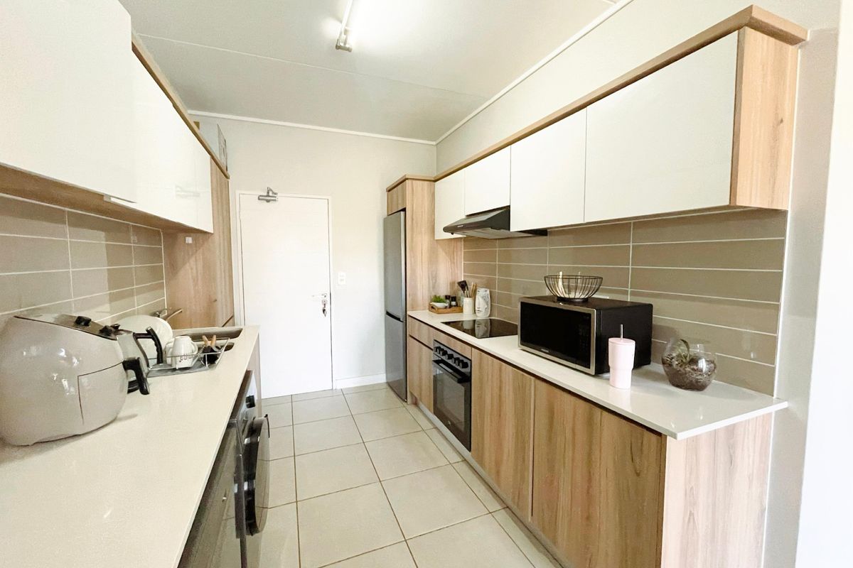 Modern galley-style kitchen featuring light wood-grain cabinetry, white countertops, tiled backsplash, and integrated appliances including an oven and hob.