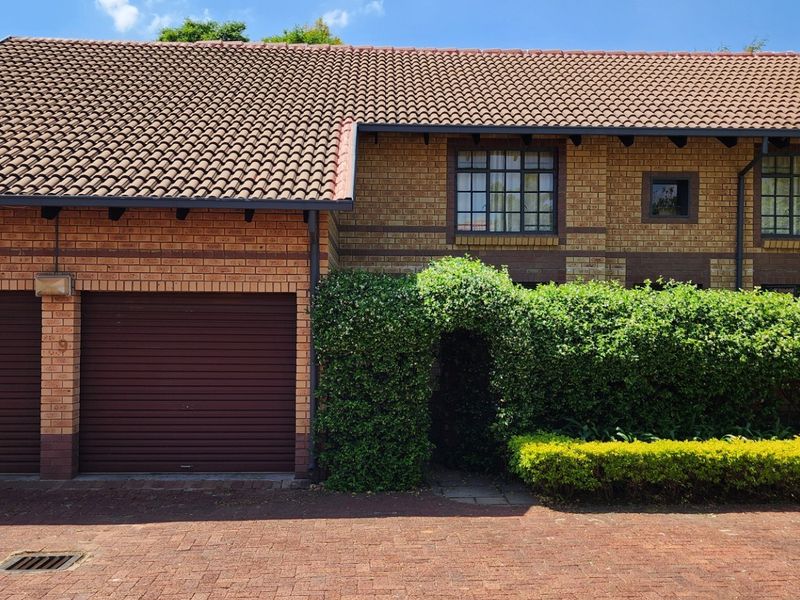 Front elevation of a townhouse featuring a double garage, brick exterior, tiled roof, and a landscaped entrance with mature hedging.